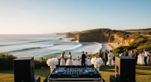 A dramatic wide-screen South Coast event scene in Kiama featuring a professional DJ setup overlooking the ocean cliffs. Subtle atmospheric lighting, guests enjoying an elegant outdoor celebration, rolling waves and rugged coastline in the background, warm golden-hour tones, premium speakers and DJ decks subtly in frame, cinematic realism capturing Kiama’s natural beauty and event ambience.